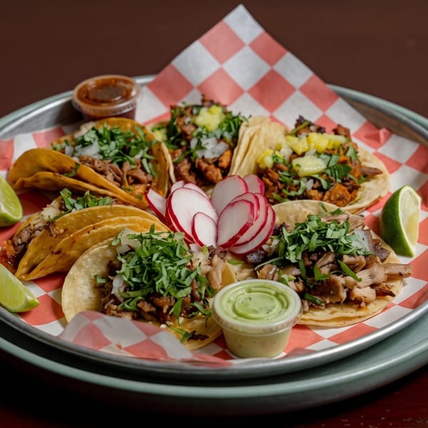 A metal tray holds various tacos with different fillings, radishes, lime wedges, and sauces.